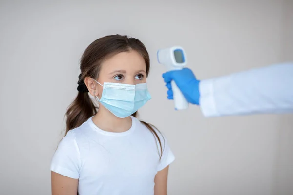 Mała dziewczynka w masce do twarzy, której temperatura została sprawdzona przez elektroniczny termometr, czekając na szczepionkę przeciw wirusowi covid-19 — Zdjęcie stockowe