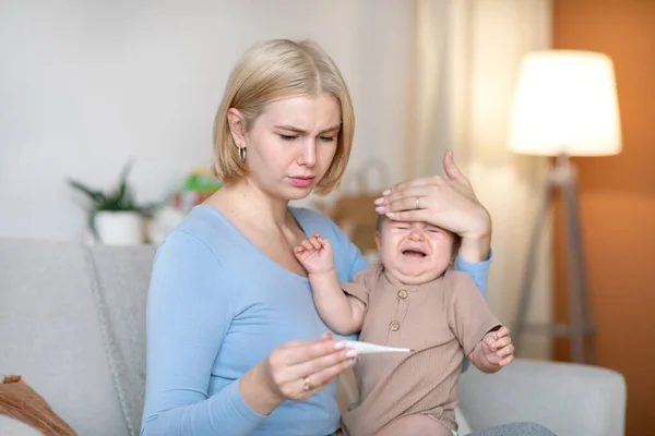 Orolig mamma tar temperaturen till sjuka gråtande barn hemma — Stockfoto