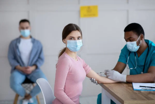 Czarny lekarz wstrzykujący domięśniowo młodej kobiecie w gabinecie lekarskim — Zdjęcie stockowe