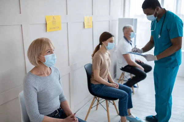 Mężczyzna lekarz rozmawia z młodą kobietą, robi notatki przed wstrzyknięciem szczepionki przeciw kovid-19 w poczekalni kliniki — Zdjęcie stockowe