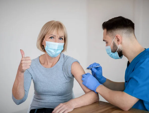 Зріла жінка в масці для обличчя, що схвалює вакцинацію проти ковідо-19, показує великий палець під час ін'єкції коронавірусної вакцини — стокове фото