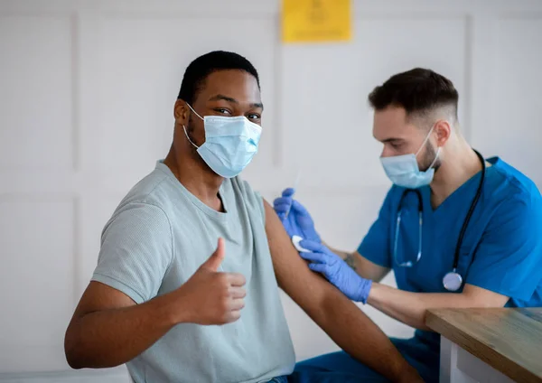 Hombre afroamericano con máscara haciendo gestos con el pulgar hacia arriba durante la vacunación contra el coronavirus, aprobando la inmunización covid-19 —  Fotos de Stock