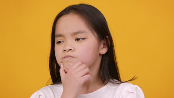 Nahaufnahme Porträt eines kleinen nachdenklichen Mädchens, das über seine Aufgabe nachdenkt, sein Kinn streichelt, orangefarbener Studiohintergrund — Stockvideo
