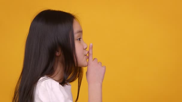 Profile portrait of little asian girl showing quiet gesture aside at free space, orange studio background — Stock Video