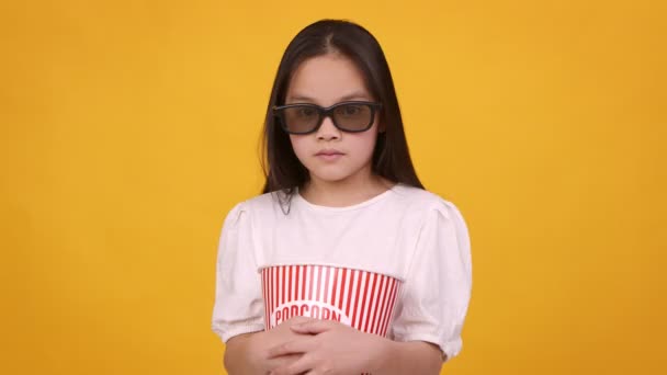 Üç boyutlu gözlüklü, elinde patlamış mısırla turuncu arka planda duran korkmuş küçük Asyalı kız. — Stok video
