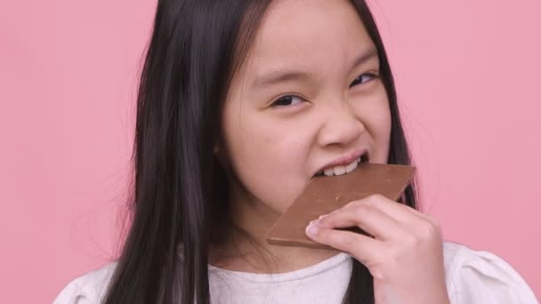 Close up retrato de menina asiática comendo chocolate, mordendo bar inteiro e apreciando, fundo de estúdio rosa — Vídeo de Stock