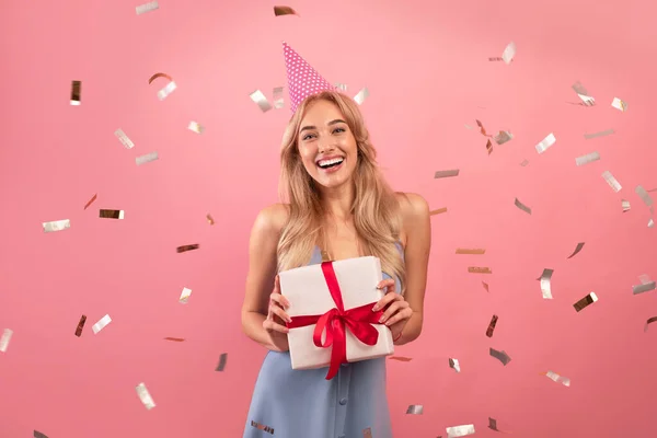 Happy young lady in birthday hat and cute dress holding gift box on pink studio background with falling confetti