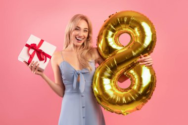 Güzel elbiseler içinde neşeli bir milenyum hanımefendisi. Pembe arka planda 8 Mart 'ta hediye paketi ve altın balon taşıyor.