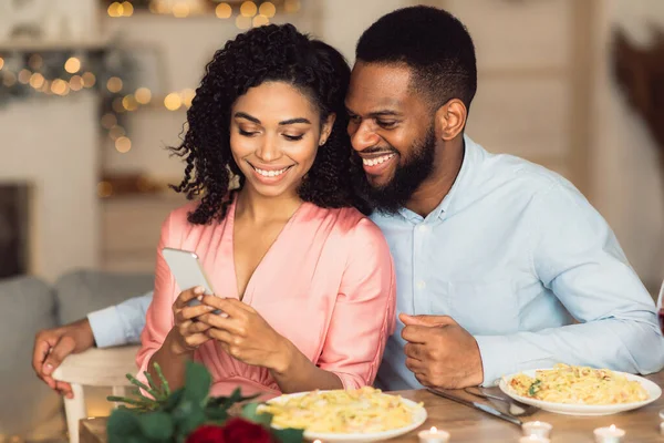 Smiling black man and woman using smartphone during romantic dinner — Stock Photo, Image