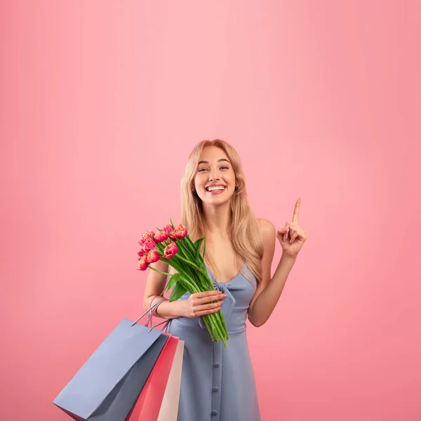 Feliz joven vistiendo precioso vestido, sosteniendo flores de primavera y bolsas de regalo, señalando el espacio vacío sobre fondo rosa — Foto de Stock
