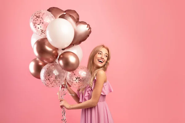 Nå er det fest. Sjarmerende blond kvinne i stilig kjole med bursdagsballonger over rosa studiobakgrunn, kopieringsrom – stockfoto