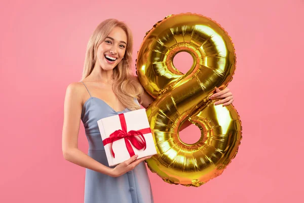 Pozitív fiatal hölgy kezében arany nyolc alakú léggömb és ajándék doboz felett rózsaszín stúdió háttér — Stock Fotó