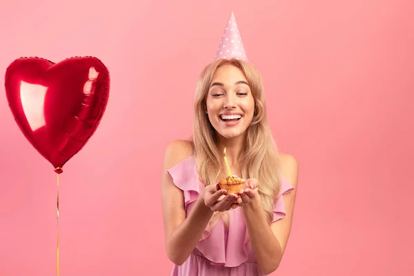 Portrett av glad blond kvinne i søt kjole som feirer og holder bursdagsmuffin med lys på rosa bakgrunn – stockfoto