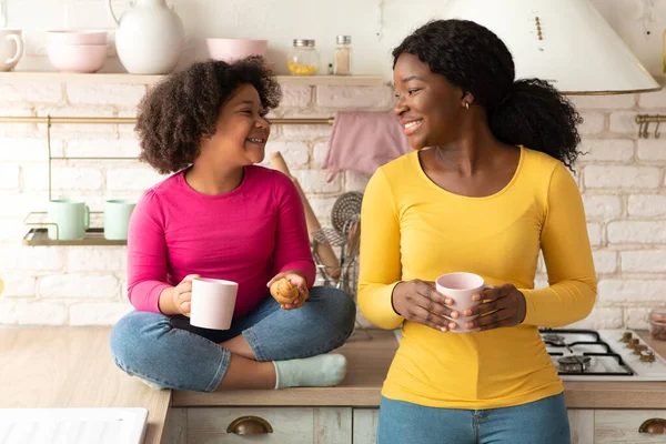 Feliz madre negra e hija pequeña hablando y comiendo bocadillos en la cocina — Foto de Stock