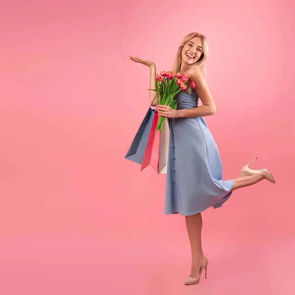 Dame blonde avec des tulipes et des sacs à provisions tenant quelque chose sur fond rose, maquette pour la conception de votre produit — Photo