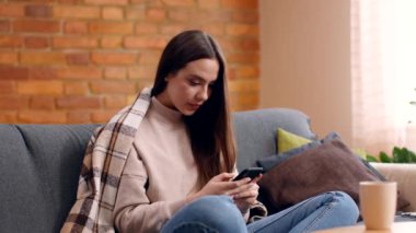 Kendini izole etmiş genç bir kadın cep telefonuyla konuşuyor, ailesiyle ya da arkadaşlarıyla iletişim kuruyor, evdeki kanepede oturuyor.