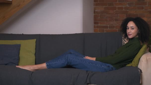 Linda mujer negra sentada en un acogedor sofá en casa, sonriendo a la cámara, vista lateral — Vídeos de Stock