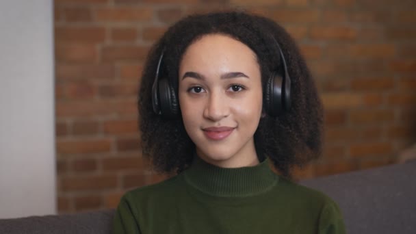 Κοντινό πορτρέτο μιας νεαρής Αφροαμερικανής γυναίκας που φοράει ασύρματα ακουστικά, απολαμβάνει το ηχητικό βιβλίο, χαμογελώντας στην κάμερα — Αρχείο Βίντεο