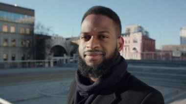 Ciddi sakallı, ceketli, boş şehir merkezinde yürüyen Afro-Amerikan bir adamın açık hava portresi.