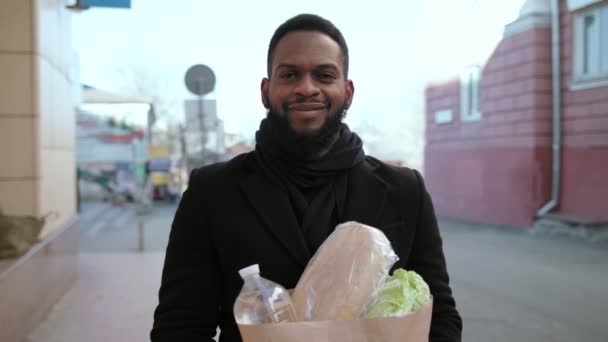Jovem negro confiante carregando saco de papel de compras com mantimentos ao ar livre, tiro de rastreamento — Vídeo de Stock