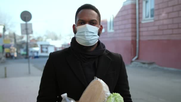 Afro-Amerikaanse man draagt een beschermend masker met papieren tas met boodschappen, die terugkomt van het winkelen — Stockvideo