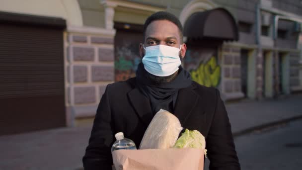 Ritratto all'aperto di uomo nero in maschera protettiva che trasporta cibo in borsa, in piedi nel centro della città, tracking shot — Video Stock