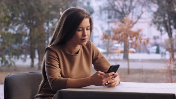 Młoda kobieta przegląda na smartfonie, siedzi sama w kawiarni przy oknie — Wideo stockowe