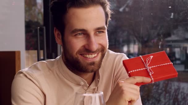 Szczęśliwy zabawny mężczyzna pokazujący prezent swojej dziewczynie i uśmiechnięty, bawiący się brwiami podczas randki w kawiarni, w zwolnionym tempie — Wideo stockowe