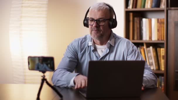Досвідчений чоловік середнього віку в навушниках записує відео блог на смартфоні, говорячи про новини бізнесу — стокове відео