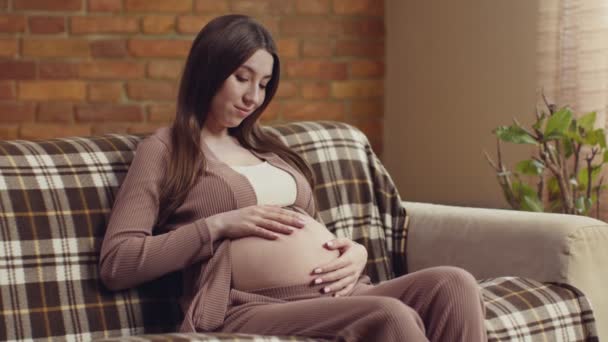 Mosolygó terhes nő ül a kanapén, és simogatja a meztelen hasát, várja, hogy találkozzon a babájával. — Stock videók
