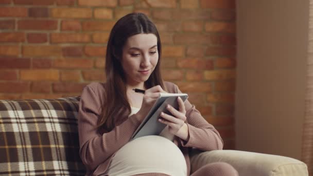 Mujer embarazada feliz escribiendo en su calendario de embarazo o preparando la lista para el parto, sentado en el sofá en casa — Vídeos de Stock