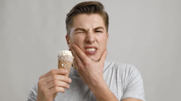 Young guy eating ice-cream cone and suffering from acute toothpain, grey studio background, close up portrait — Stock Video