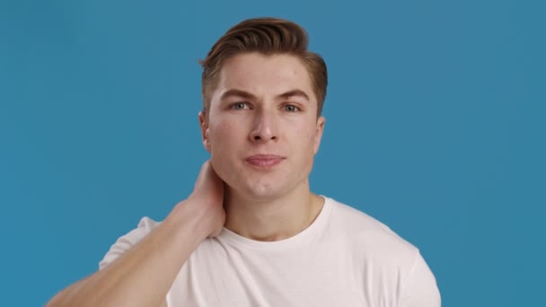 Junger Mann massiert seinen schmerzenden Hals, blauer Hintergrund, Zeitlupe — Stockvideo