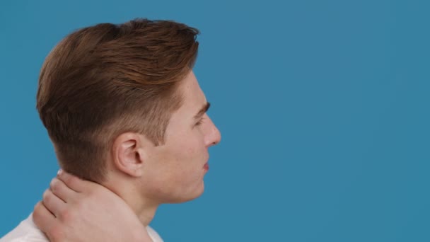 Молодой человек страдает от боли в шее, массируя ее, портрет профиля, синий студийный фон — стоковое видео