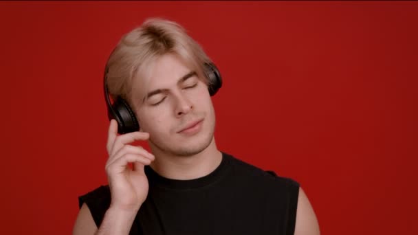 Studio portret młodego blondyna słuchającego muzyki w słuchawkach i tańczącego, czerwone tło — Wideo stockowe