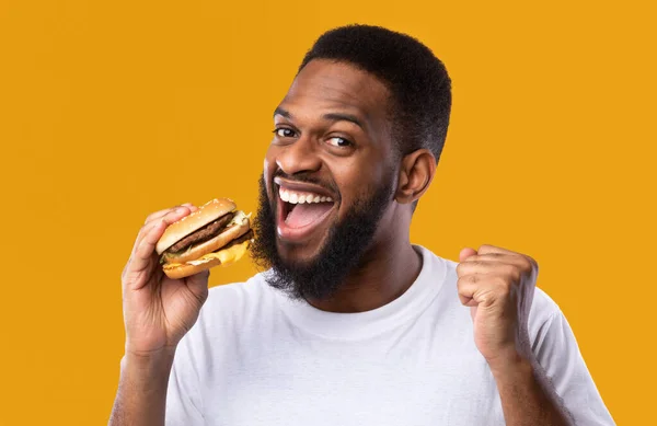 Aufgeregter Afrikaner isst Burger und gestikuliert vor gelbem Hintergrund — Stockfoto