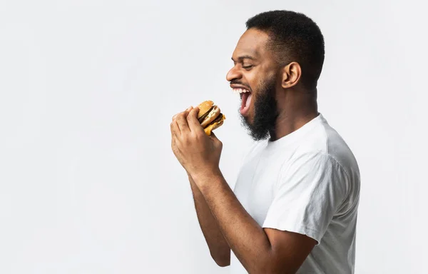 Vista lateral do homem negro faminto comer hambúrguer, fundo branco — Fotografia de Stock