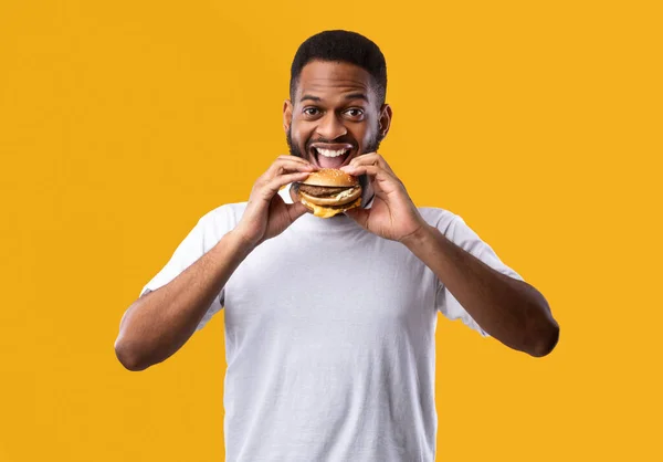 Fröhlicher hungriger afrikanischer Kerl isst Burger auf gelbem Hintergrund — Stockfoto