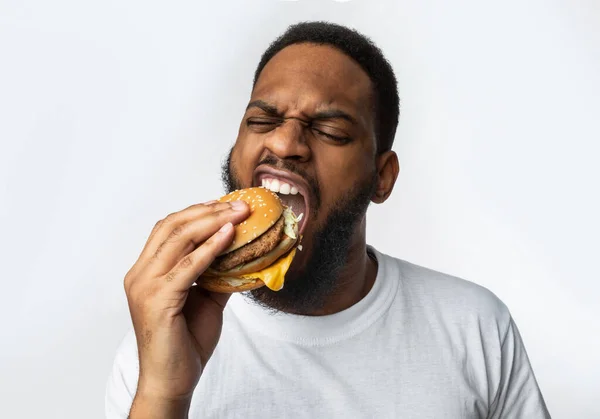 Homem africano mordendo hambúrguer desfrutando de junk food sobre fundo branco — Fotografia de Stock