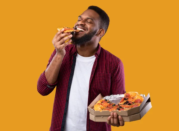 Jovem africano desfrutando de pizza posando com caixa, fundo amarelo — Fotografia de Stock