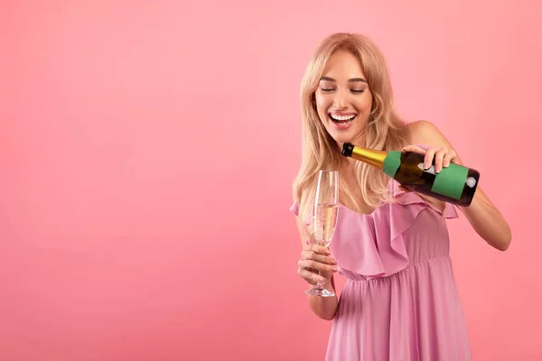 Senhora loira alegre derramando champanhe em vidro sobre fundo de estúdio rosa, espaço livre. Conceito festivo — Fotografia de Stock