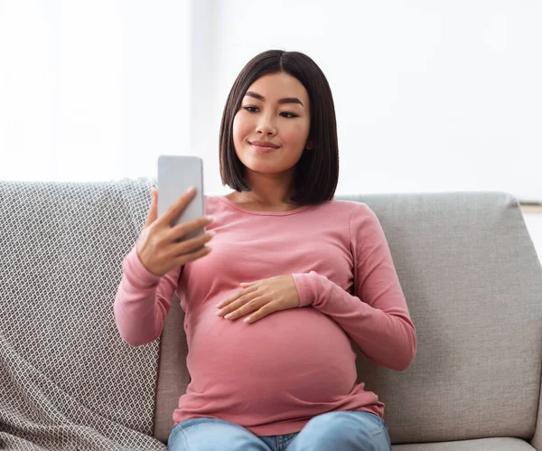 Incinta cinese signora utilizzando telefono making selfie seduta a casa — Foto Stock