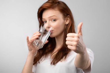 Kızıl saçlı, su içen genç bayan Gri Arkaplan 'a başparmak hareketi yapıyor.