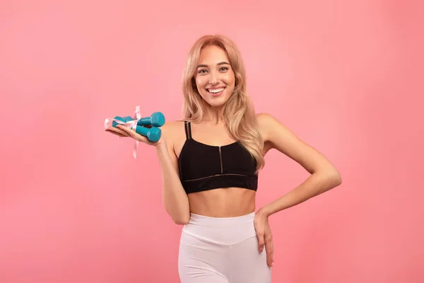 Happy fit young lady holding dumbbells tied by ribbon on pink studio background. Active lifestyle and wellness concept — Stock Photo, Image