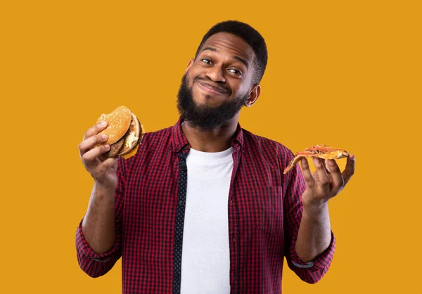 Hombre africano sosteniendo hamburguesa y rebanada de pizza, fondo amarillo — Foto de Stock