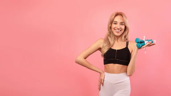 Preciosa mujer rubia deportiva sosteniendo mancuernas con cinta de regalo sobre fondo rosa, diseño de pancarta con espacio para copiar — Foto de Stock