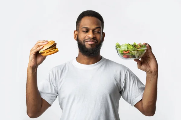 Chico africano sosteniendo hamburguesa y ensalada eligiendo dieta, fondo blanco —  Fotos de Stock