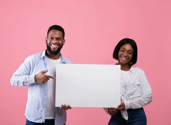 Joyful black couple holding and pointing at empty advertising board with copyspace on pink background, mockup for design