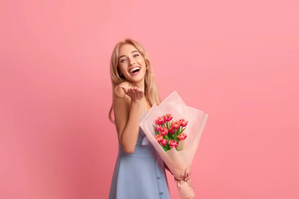 Jublende ung dame med nydelige vårblomster som ler og ser på kamera på rosa studiobakgrunn – stockfoto