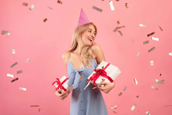 Heureuse dame millénaire tenant des boîtes-cadeaux enveloppées, portant un chapeau d'anniversaire sur fond de studio rose avec des confettis tombants — Photo